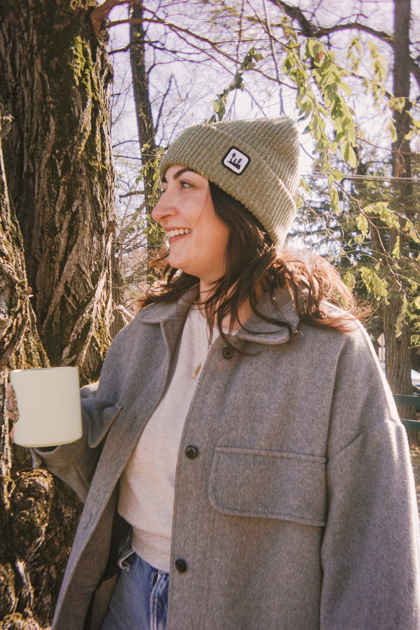 Person wearing a gray coat and green woosah beanie holding a white mug outdoors.